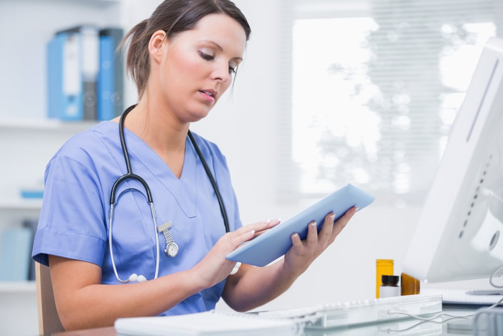 Side view of young female surgeon using digital tablet in front of computer at desk in clinic-1